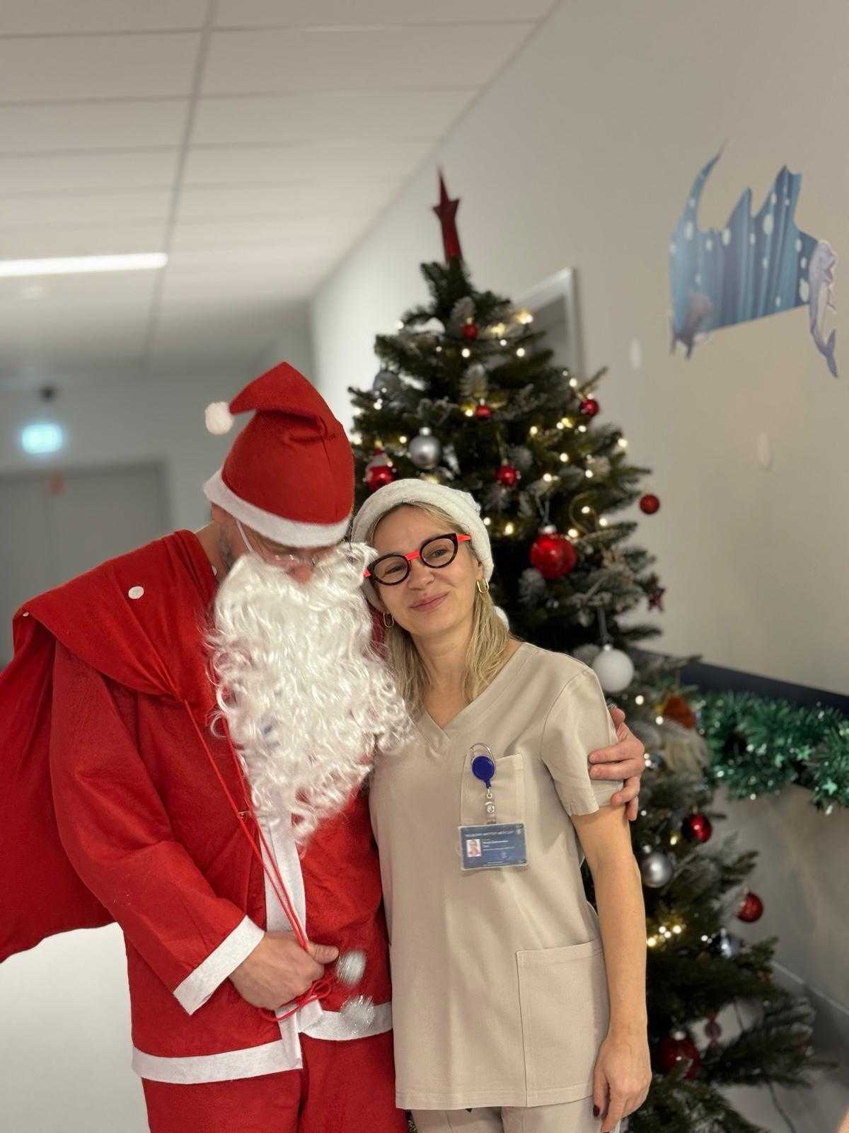 Święty Mikołaj pozuje na korytarzu szpitalnym z pracowniczką szpitala. Za nimi stoi udekorowana choinka.
