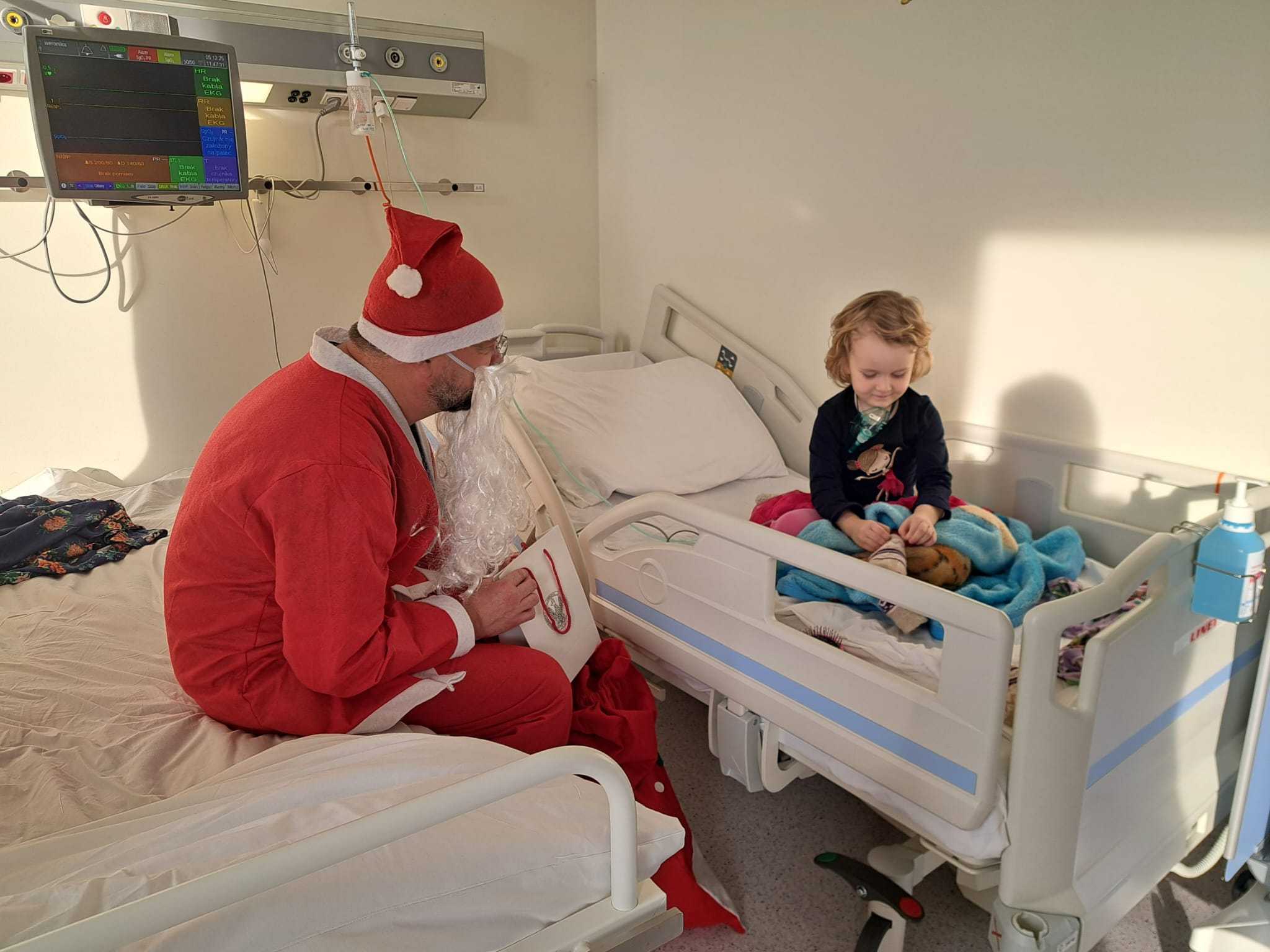 Święty Mikołaj siedzi na łóżku szpitalnym naprzeciw niego na drugim łóżku siedzi małą dziewczynka i ogląda prezenty.