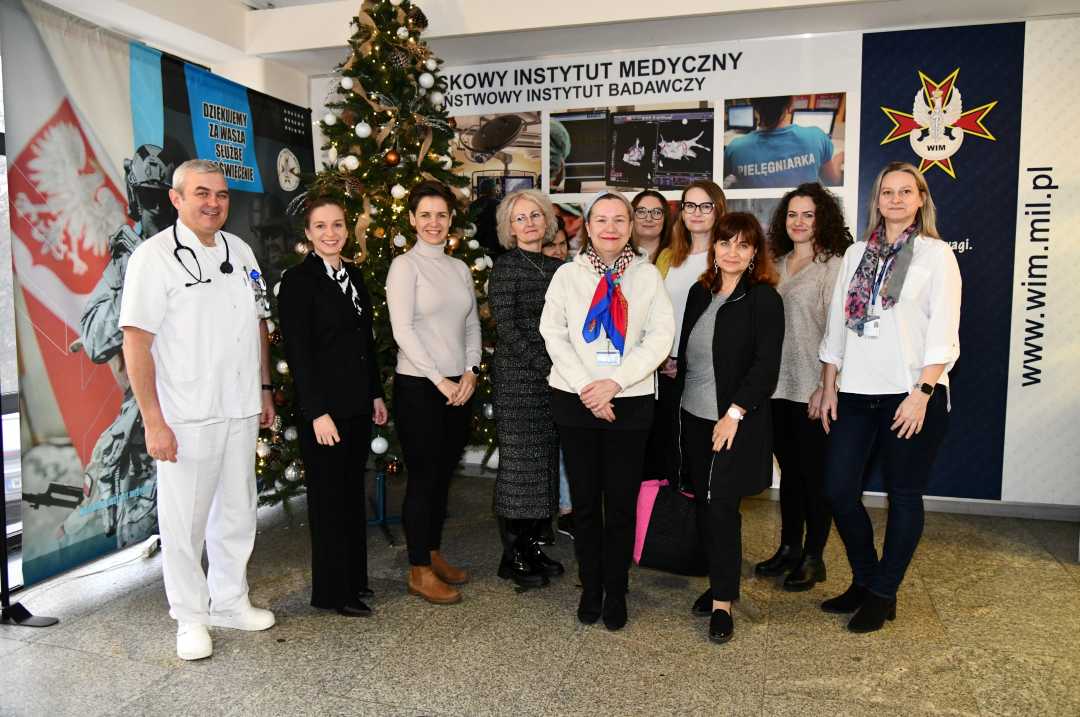 Organizatorzy i uczestnicy spotkania pozują do zdjęcia na tle ścianki Centrum Leczenia Weterana. Z tyłu za pozującymi stoi choinka bożonarodzeniowa.