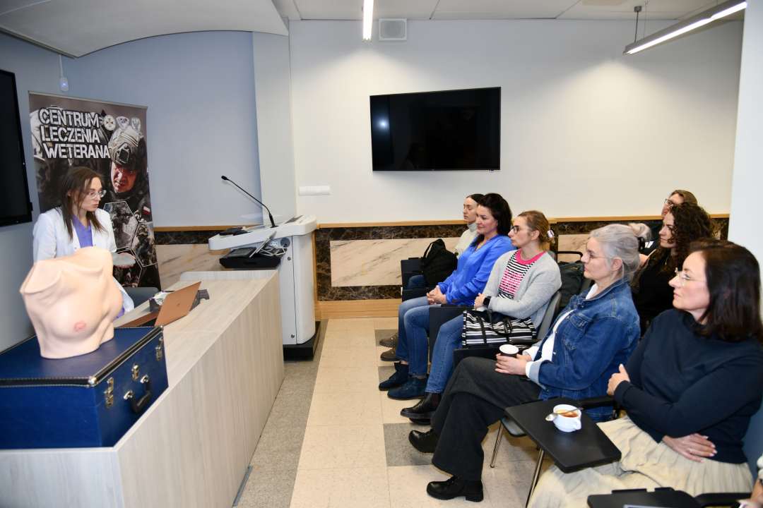 Sala wykładowa. Lekarka prowadząca prelekcję obok niej na katedrze stoi fantom kobiecej klatki piersiowej. Na przeciwko niej siedzące słuchaczki.