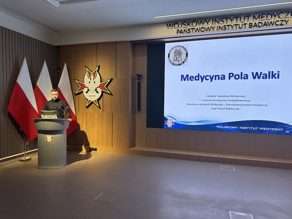 Prelegent przy mównicy podczas konferencji, za nim na wyświetlaczu prezentacja Medycyna Pola Walki.