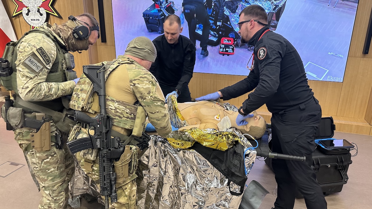 Pokaz ratownictwa medycyny pola walki na auli WIM. Ratownicy przy fantomie poszkodowanego żołnierza. Na ścianach dwa duże monitory wyświetla relację z pokazu.