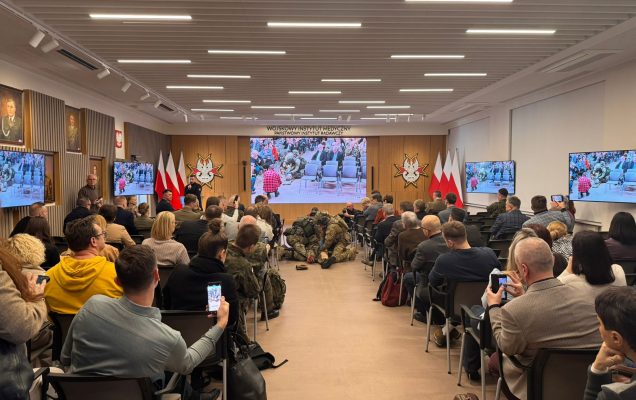 Ujęcie z tyłu sali. Uczestnicy konferencji oglądają pokaz ratownictwa medycyny pola walki.
