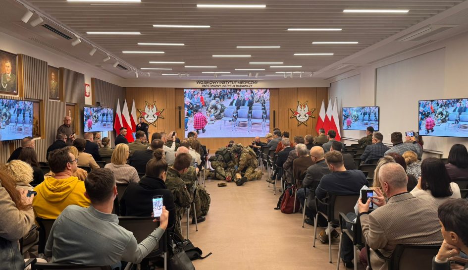 Ujęcie z tyłu sali. Uczestnicy konferencji oglądają pokaz ratownictwa medycyny pola walki.