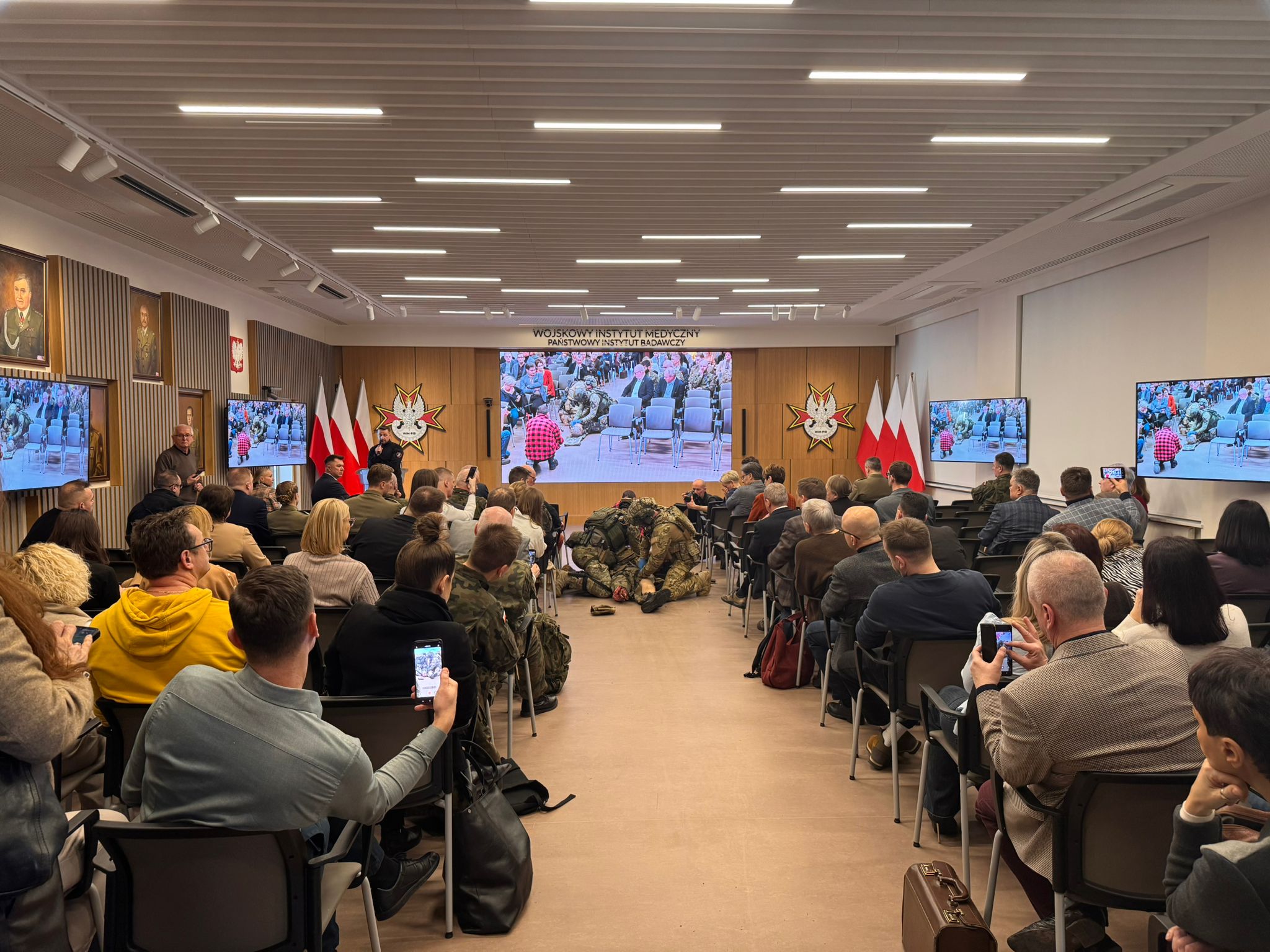Ujęcie z tyłu sali. Uczestnicy konferencji oglądają pokaz ratownictwa medycyny pola walki.