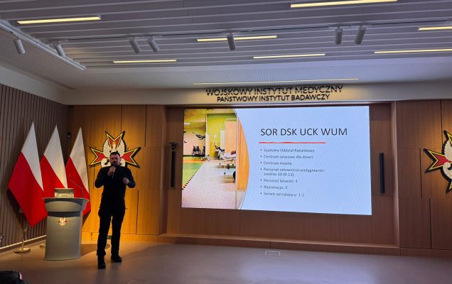 Prelegent przy mównicy. Za nim na dużym monitorze slajd z prezentacji.