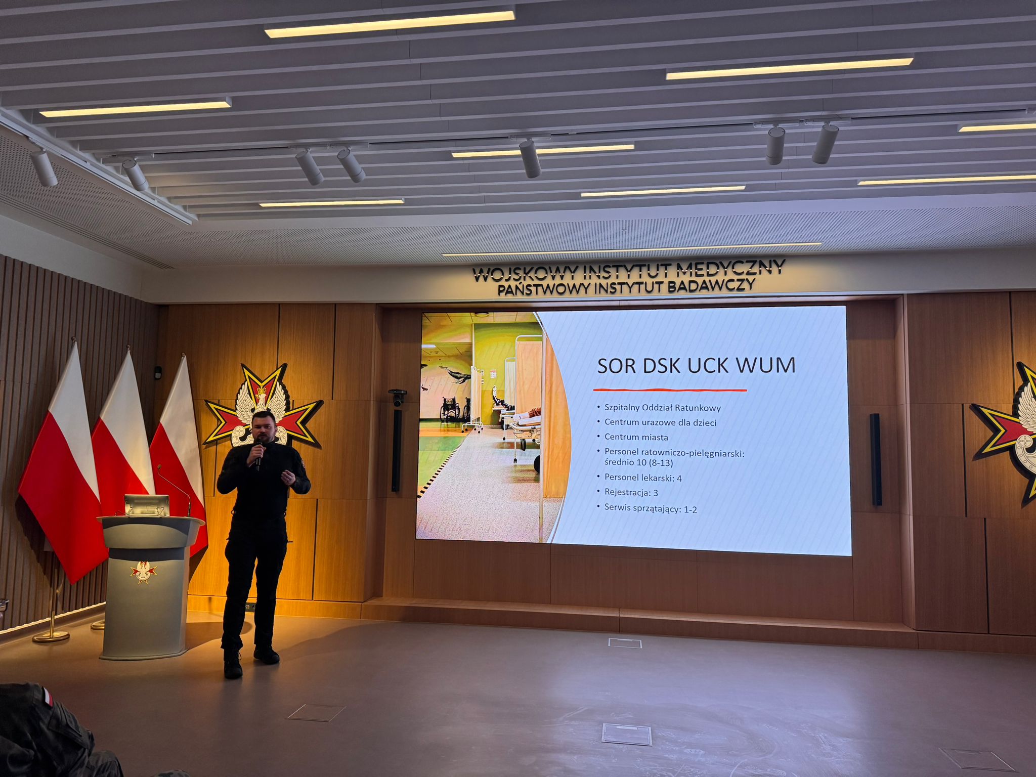 Prelegent przy mównicy. Za nim na dużym monitorze slajd z prezentacji.