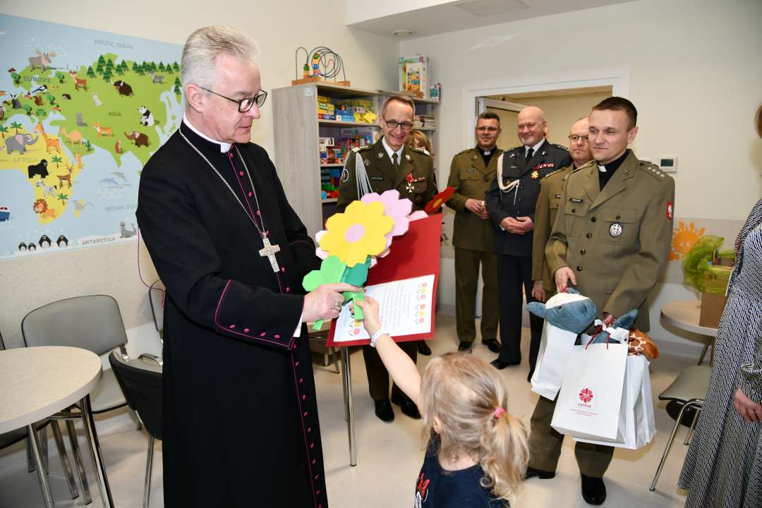 Sala szpitalna. Mała dziewczynka wręcza biskupowi papierowy kwiat.