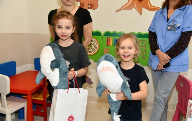 Dwie małe uśmiechnięte dziewczynki z maskotkami rekina i torbami z prezentami od biskupa.