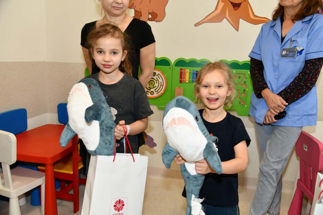 Dwie małe uśmiechnięte dziewczynki z maskotkami rekina i torbami z prezentami od biskupa.
