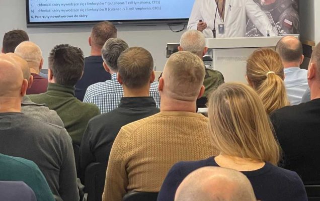 Doktor Karol Pniewski przy mównicy podczas wykładu, za nim ekran z wyświetlaną prezentacją.