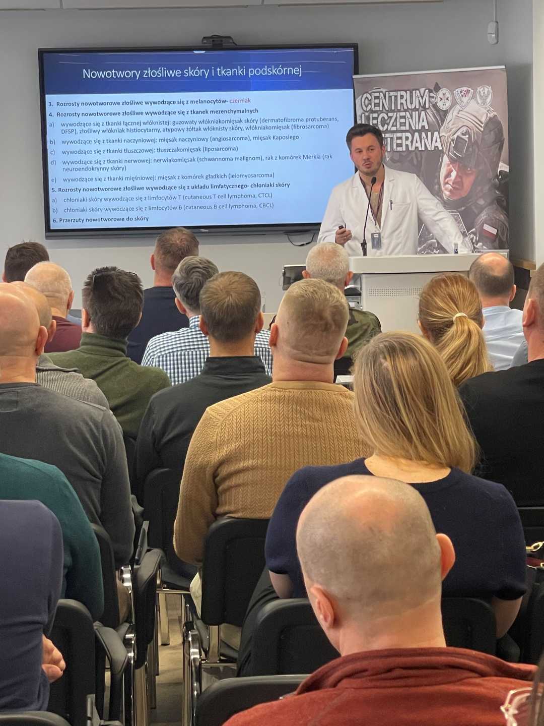 Doktor Karol Pniewski przy mównicy podczas wykładu, za nim ekran z wyświetlaną prezentacją.