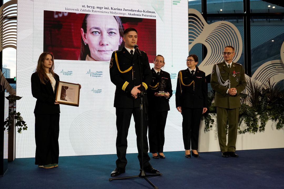 Członkowie Akademii Pożarniczej na scenie. Za nimi na ekranie zdjęcie Kariny Jarosławskiej-Kolman.