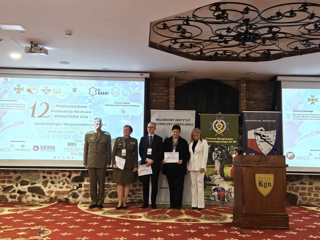 Stojący wykładowcy sesji otwierającej w sali konferencyjnej, za nimi wyświetlona plansza konferencji.