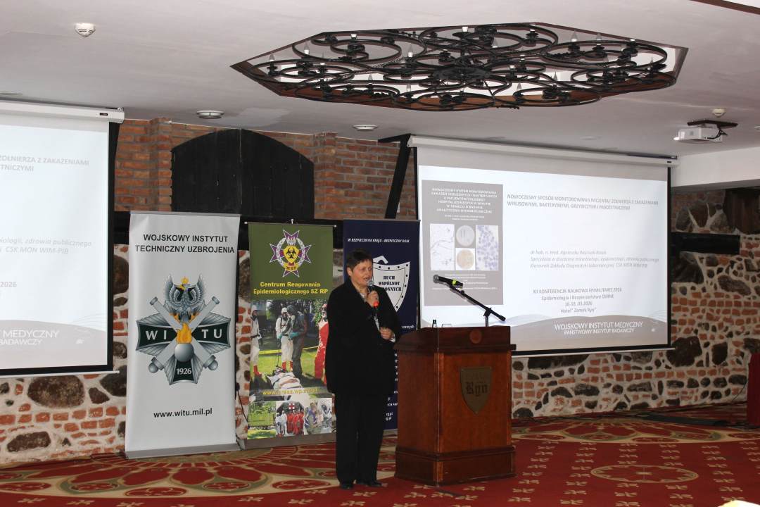 Agnieszka Woźniak przy mównicy w sali konferencyjnej, za nią wyświetlona plansza konferencji.
