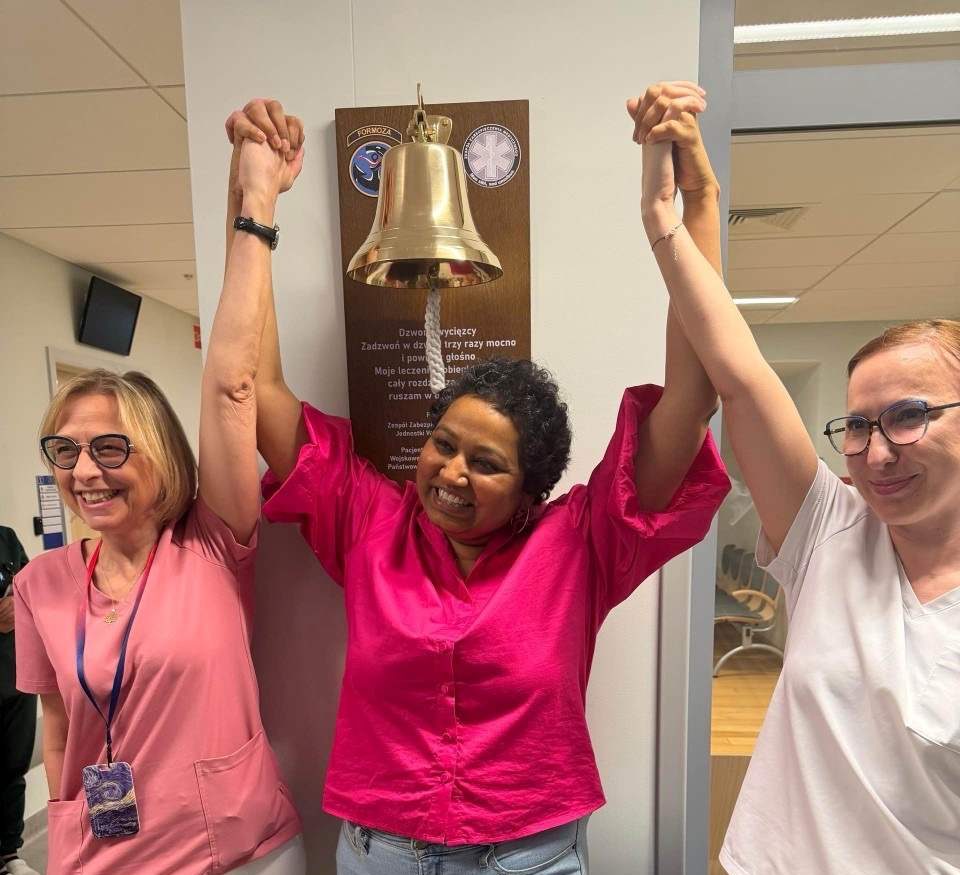 Uśmiechnięta pacjentka trzyma się za ręce w geście zwycięstwa z Renatą Duchnowską i pielęgniarką przy dzwonie zwycięscy.