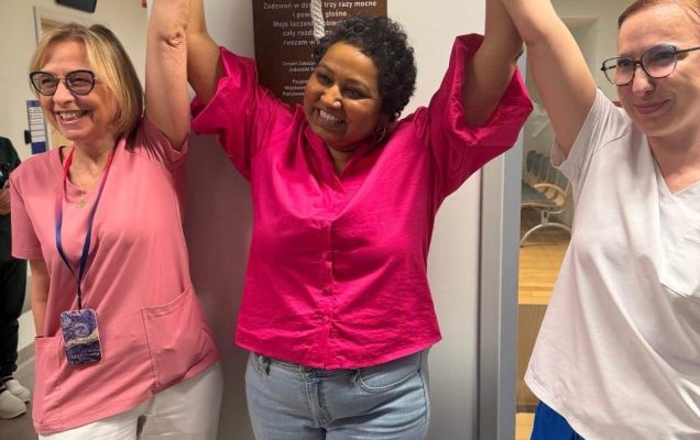 Uśmiechnięta pacjentka trzyma się za ręce w geście zwycięstwa z Renatą Duchnowską i pielęgniarką przy dzwonie zwycięscy.