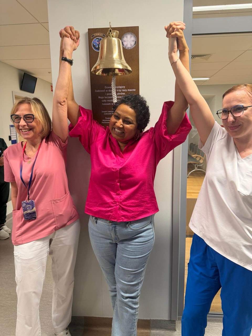 Uśmiechnięta pacjentka trzyma się za ręce w geście zwycięstwa z Renatą Duchnowską i pielęgniarką przy dzwonie zwycięscy.