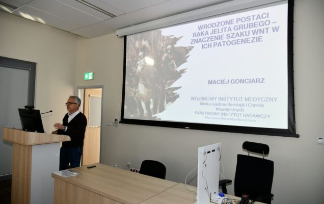 Maciej Gonciarz przy mównicy na sali WIM. Za nim wyświetlony slajd z prezentacji.