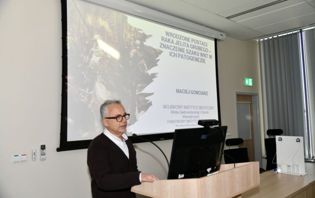 Maciej Gonciarz przy mównicy na sali WIM. Za nim wyświetlony slajd z prezentacji.