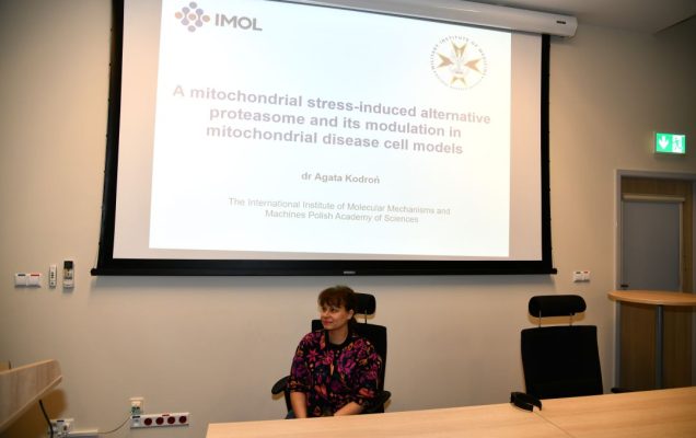 Agata Kodroń przy biurku, za nią tablica z prezentacją.