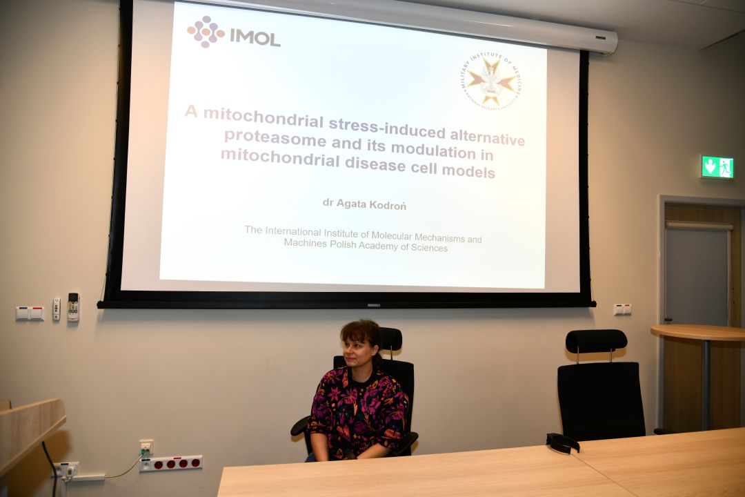 Agata Kodroń przy biurku, za nią tablica z prezentacją.