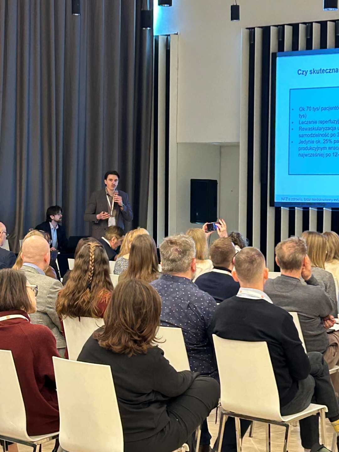 Ujęcie z tyłu sali słuchaczy i prelegenta podczas wykładu na sali konferencyjnej.