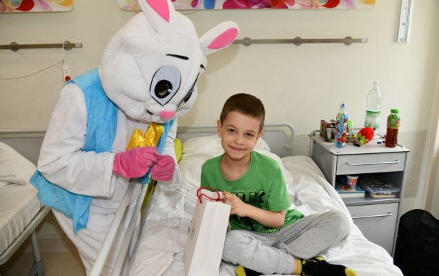 Zajączek wielkanocny w sali szpitalnej z młodym pacjentem siedzącym na łóżku trzymającym torbę z prezentem.