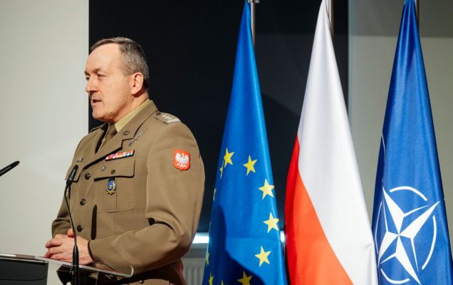 Prelegent przy mównicy. Za jego plecami flagi UE, Polski, NATO.