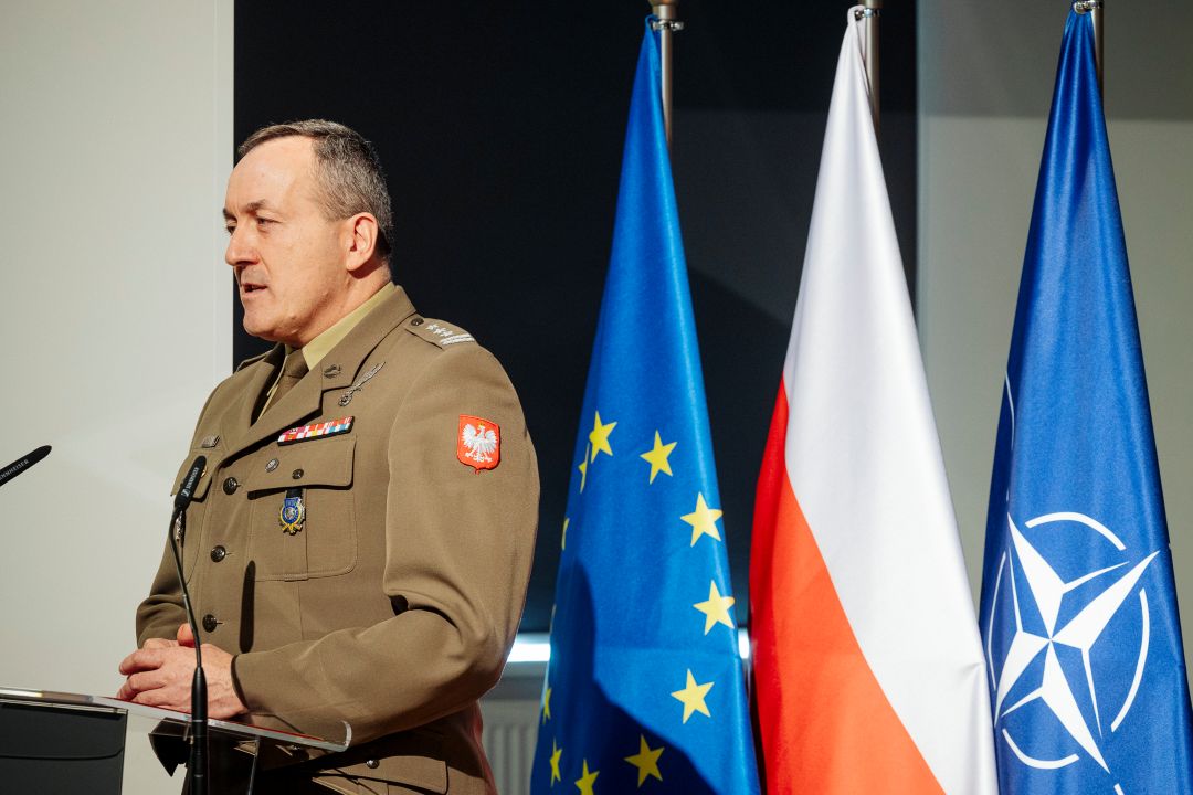 Prelegent przy mównicy. Za jego plecami flagi UE, Polski, NATO.