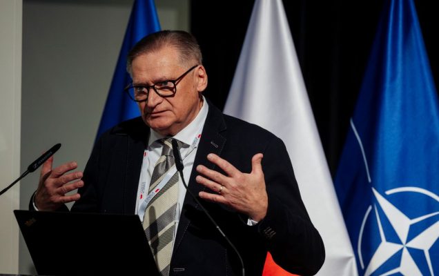 Prelegent przy mównicy. Za jego plecami flagi UE, Polski, NATO.