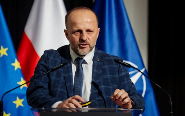 Prelegent przy mównicy. Za jego plecami flagi UE, Polski, NATO.