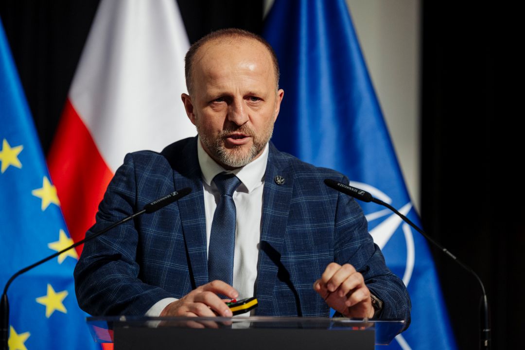 Prelegent przy mównicy. Za jego plecami flagi UE, Polski, NATO.