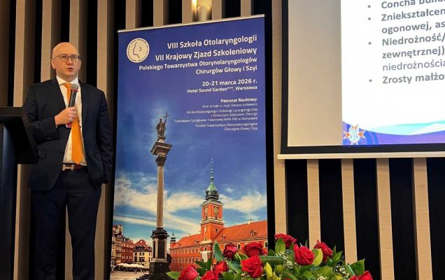 Prelegent na scenie podczas prezentacji. Mężczyzna trzyma mikrofon w ręku i patrzy na prezentację na ekranie. Przed nim kosz z bukietem kwiatów.