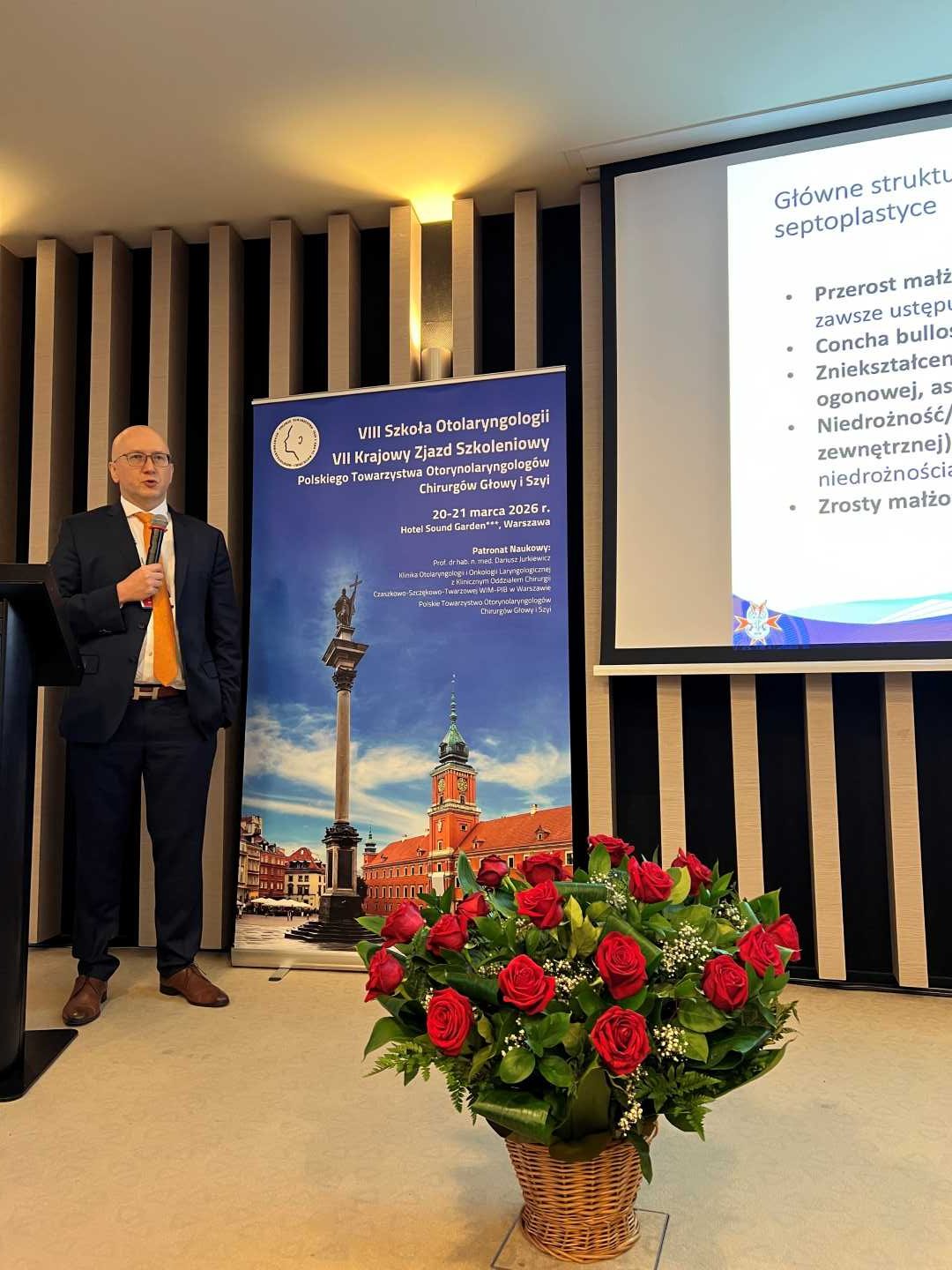 Prelegent na scenie podczas prezentacji. Mężczyzna trzyma mikrofon w ręku i patrzy na prezentację na ekranie. Przed nim kosz z bukietem kwiatów.