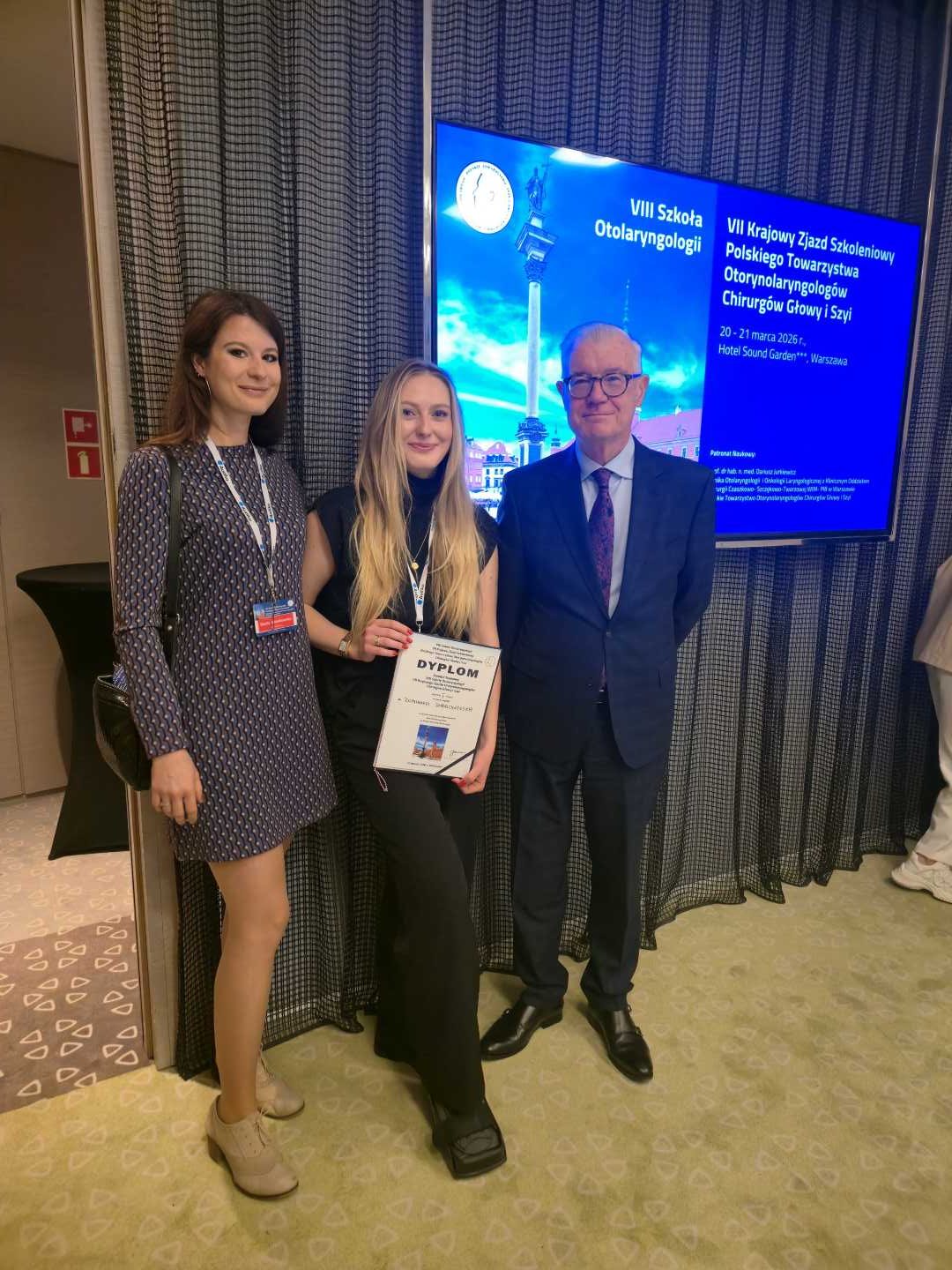 Dwie młode kobiety ze starszym mężczyzną. Jedna z kobiet trzyma w rękach dyplom. Za nimi ekran z wyświetloną prezentacją.
