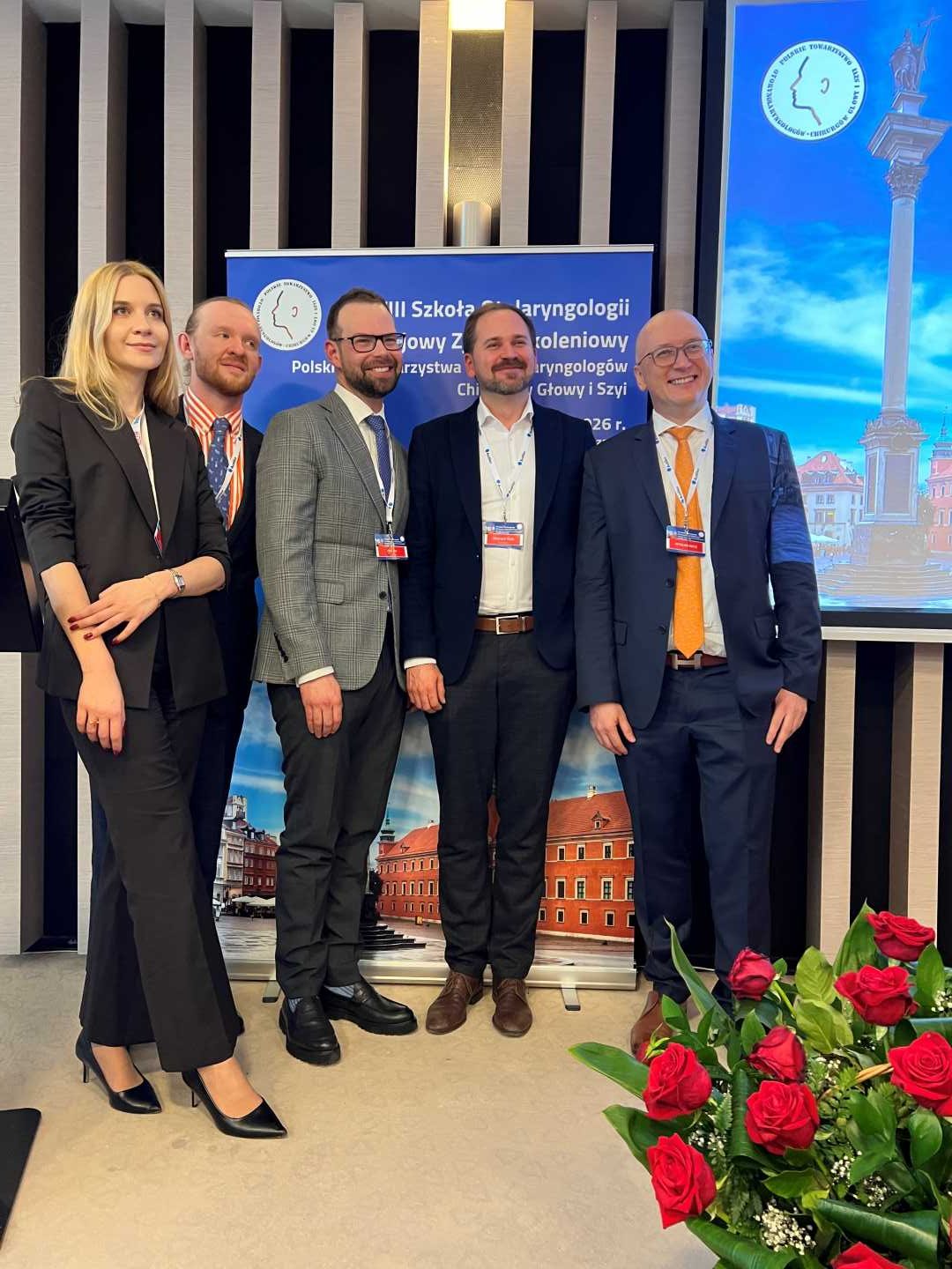 Zdjęcie grupowe prelegentów. Na scenie stoi młoda kobieta i czterech mężczyzn pozują do zdjęcia na tle ścianki z wydarzeniem.