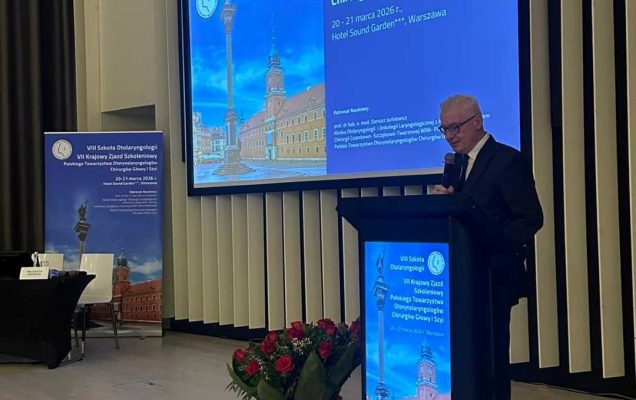 Prelegent przy mównicy. Za jego plecami na ekranie wyświetlona prezentacja.