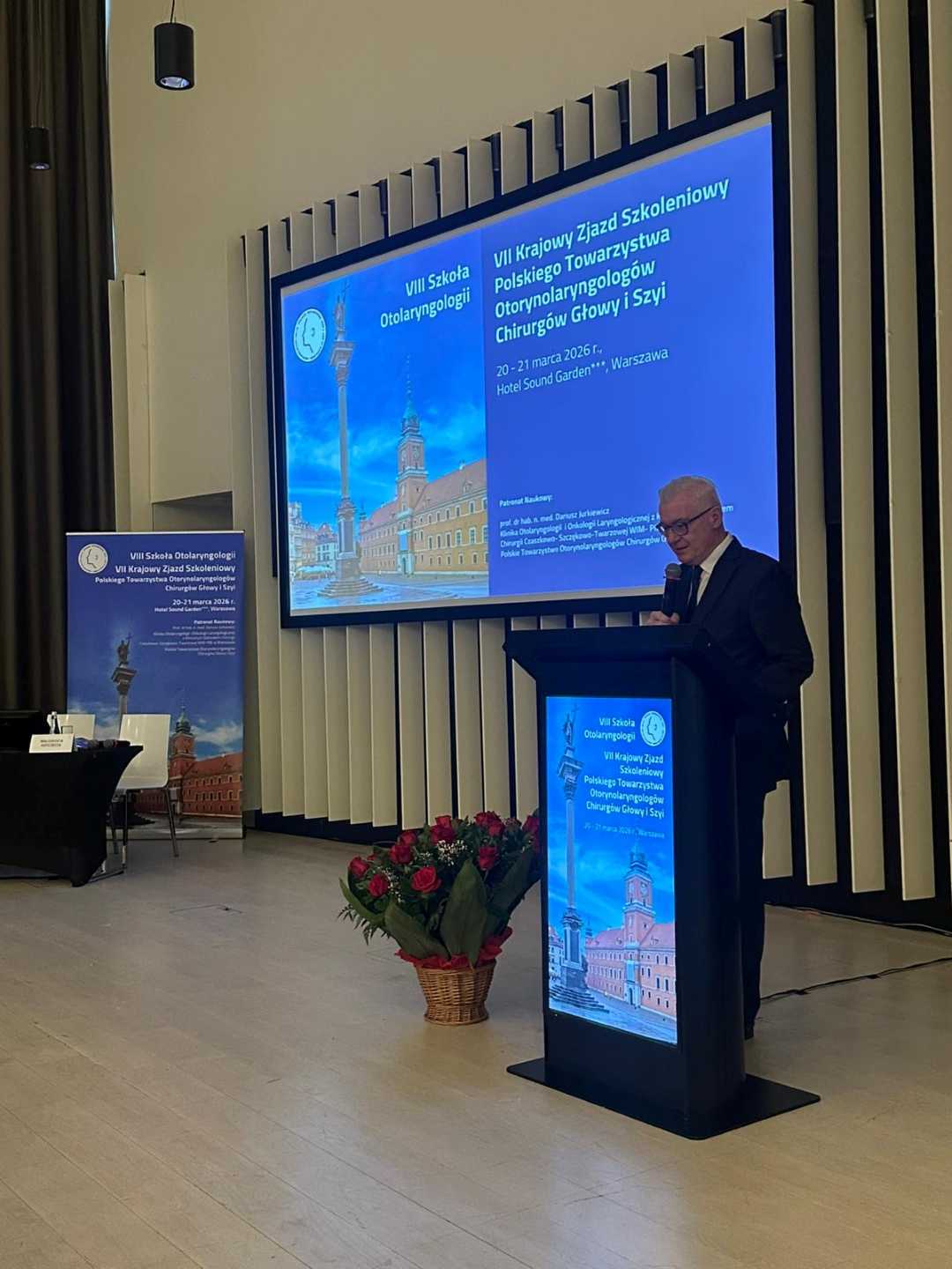 Prelegent przy mównicy. Za jego plecami na ekranie wyświetlona prezentacja.