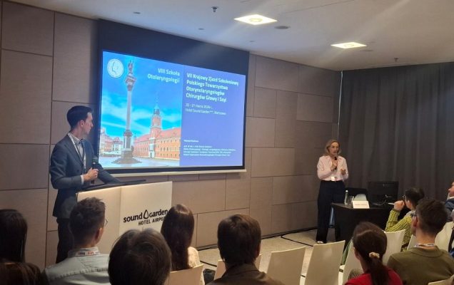 Prelegent przy mównicy. Obok prelegentka z mikrofonem. Za ich plecami na ekranie wyświetlona prezentacja.