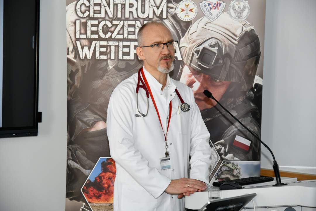 Piotr Dąbrowiecki w sali wykładowej podczas wykładu. Za jego plecami ścianka reklamowa Centrum leczenia Weterana z żołnierzem podczas ćwiczeń na poligonie.