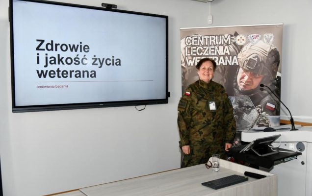 Barbara Betiuk w sali wykładowej. Obok niej na ekranie slajd z prezentacji z napisem Zdrowie i jakość życia weterana. Za jej plecami ścianka reklamowa Centrum leczenia Weterana z żołnierzem podczas ćwiczeń na poligonie.
