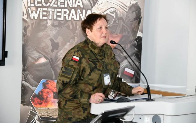 Barbara Betiuk w sali wykładowej. Za jej plecami ścianka reklamowa Centrum leczenia Weterana z żołnierzem podczas ćwiczeń na poligonie.