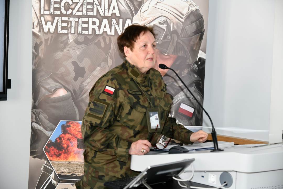 Barbara Betiuk w sali wykładowej. Za jej plecami ścianka reklamowa Centrum leczenia Weterana z żołnierzem podczas ćwiczeń na poligonie.