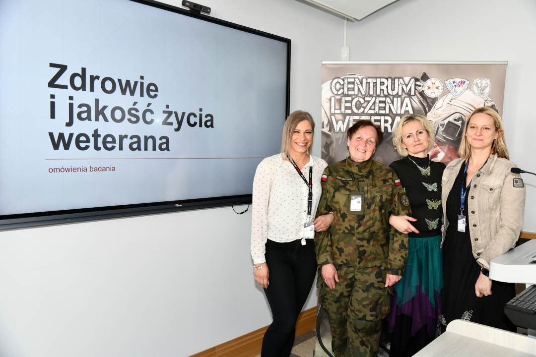 Barbara Betiuk, Marta Mnitowska, Marta Machunik, Karolina Wyczółkowska w sali wykładowej. Obok nich na ekranie slajd z prezentacji z napisem Zdrowie i jakość życia weterana. Za ich plecami ścianka reklamowa Centrum leczenia Weterana z żołnierzem podczas ćwiczeń na poligonie.