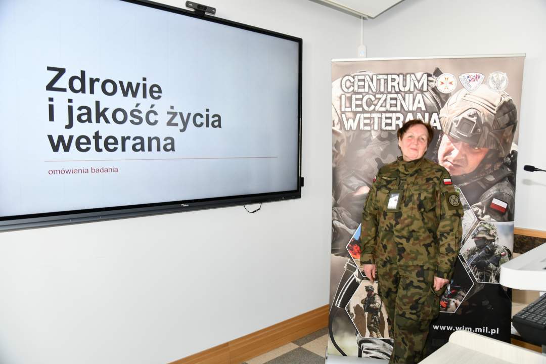 Barbara Betiuk w sali wykładowej. Obok niej na ekranie slajd z prezentacji z napisem Zdrowie i jakość życia weterana. Za jej plecami ścianka reklamowa Centrum leczenia Weterana z żołnierzem podczas ćwiczeń na poligonie.