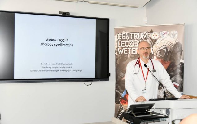 Piotr Dąbrowiecki w sali wykładowej. Obok niego na ekranie slajd z prezentacji z napisem Astma i POCHP choroby cywilizacyjne. Za jego plecami ścianka reklamowa Centrum leczenia Weterana z żołnierzem podczas ćwiczeń na poligonie.