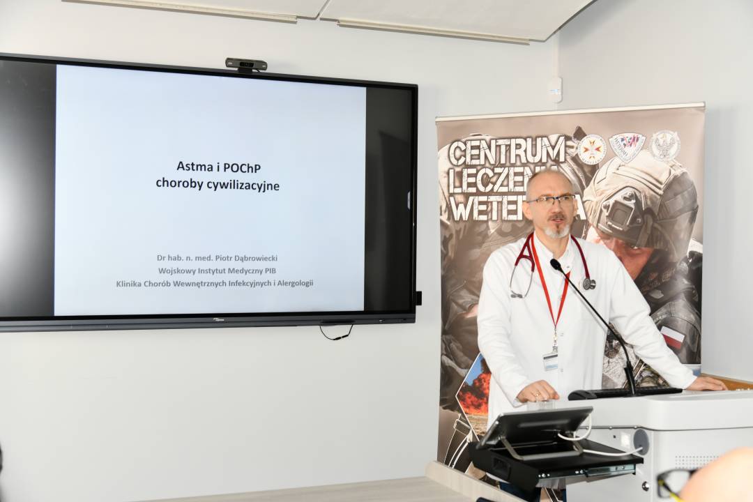 Piotr Dąbrowiecki w sali wykładowej. Obok niego na ekranie slajd z prezentacji z napisem Astma i POCHP choroby cywilizacyjne. Za jego plecami ścianka reklamowa Centrum leczenia Weterana z żołnierzem podczas ćwiczeń na poligonie.
