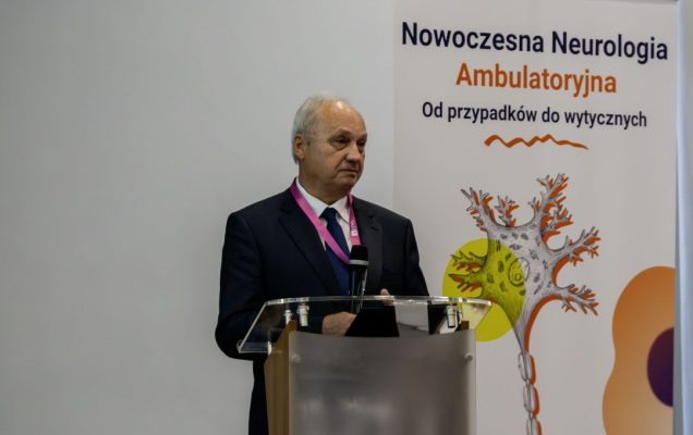 Prelegent przy mównicy podczas konferencji, za nim ścianka reklamująca konferencję.