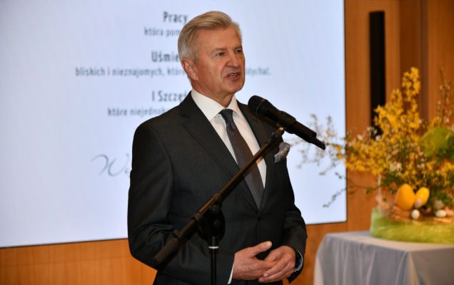 Stanisław Wziątek przy mikrofonie, za nim na ekranie wyświetlone życzenia wielkanocne.